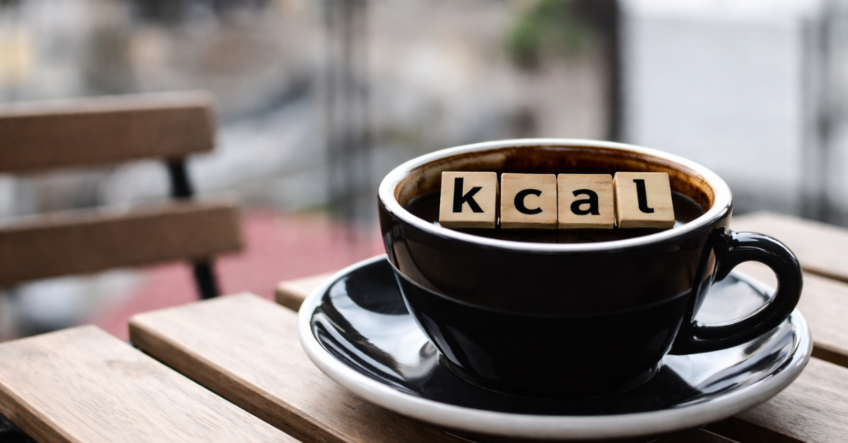 Xícara de café preto em uma mesa de madeira, com letras de madeira formando a palavra “kcal” sobre o café, em um ambiente externo desfocado.